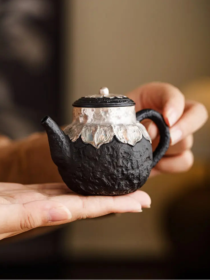 Hand holding a compact black zisha teapot with silver-finished lotus details
