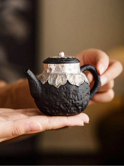 Hand holding a compact black zisha teapot with silver-finished lotus details
