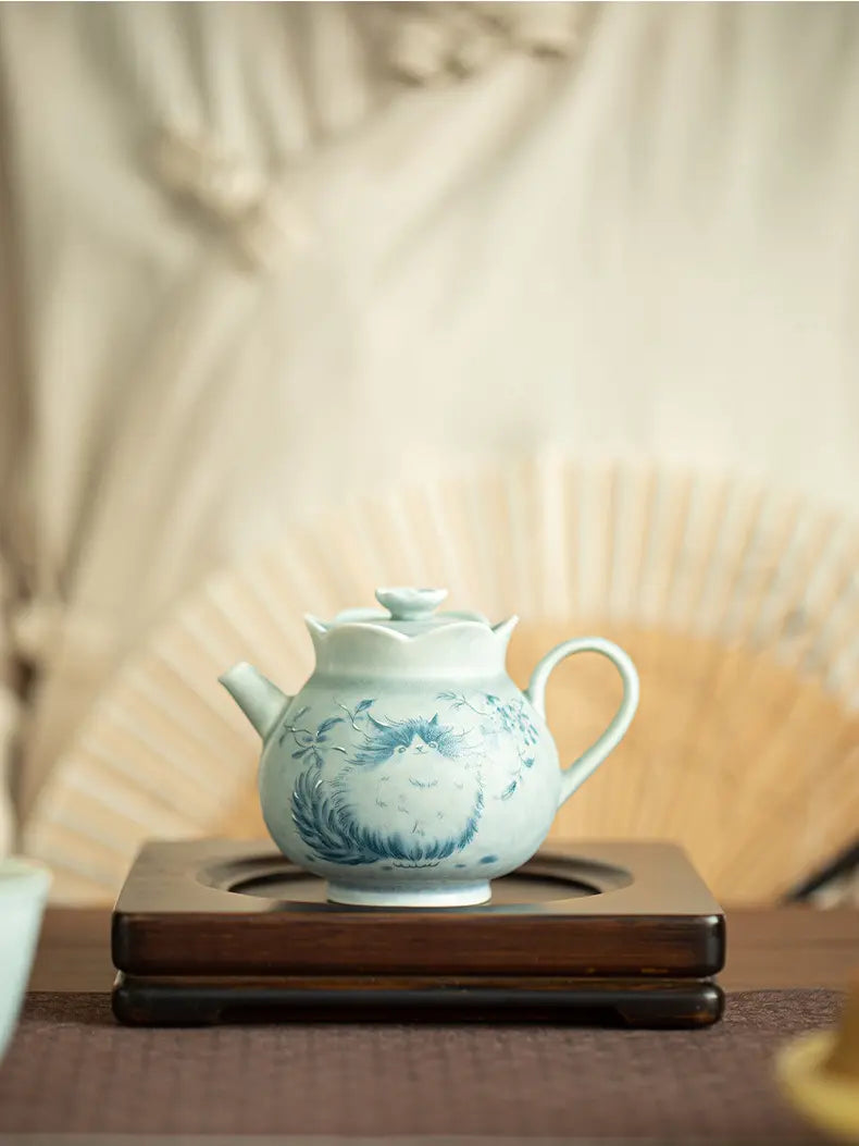 Pale green cat ceramic teapot on a dark wooden stand with a folding fan backdrop
