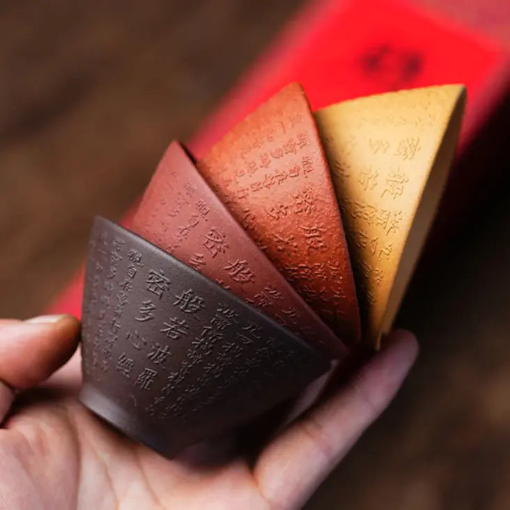 Hand holding four earth-tone Heart Sutra zisha tea cups arranged in a fan