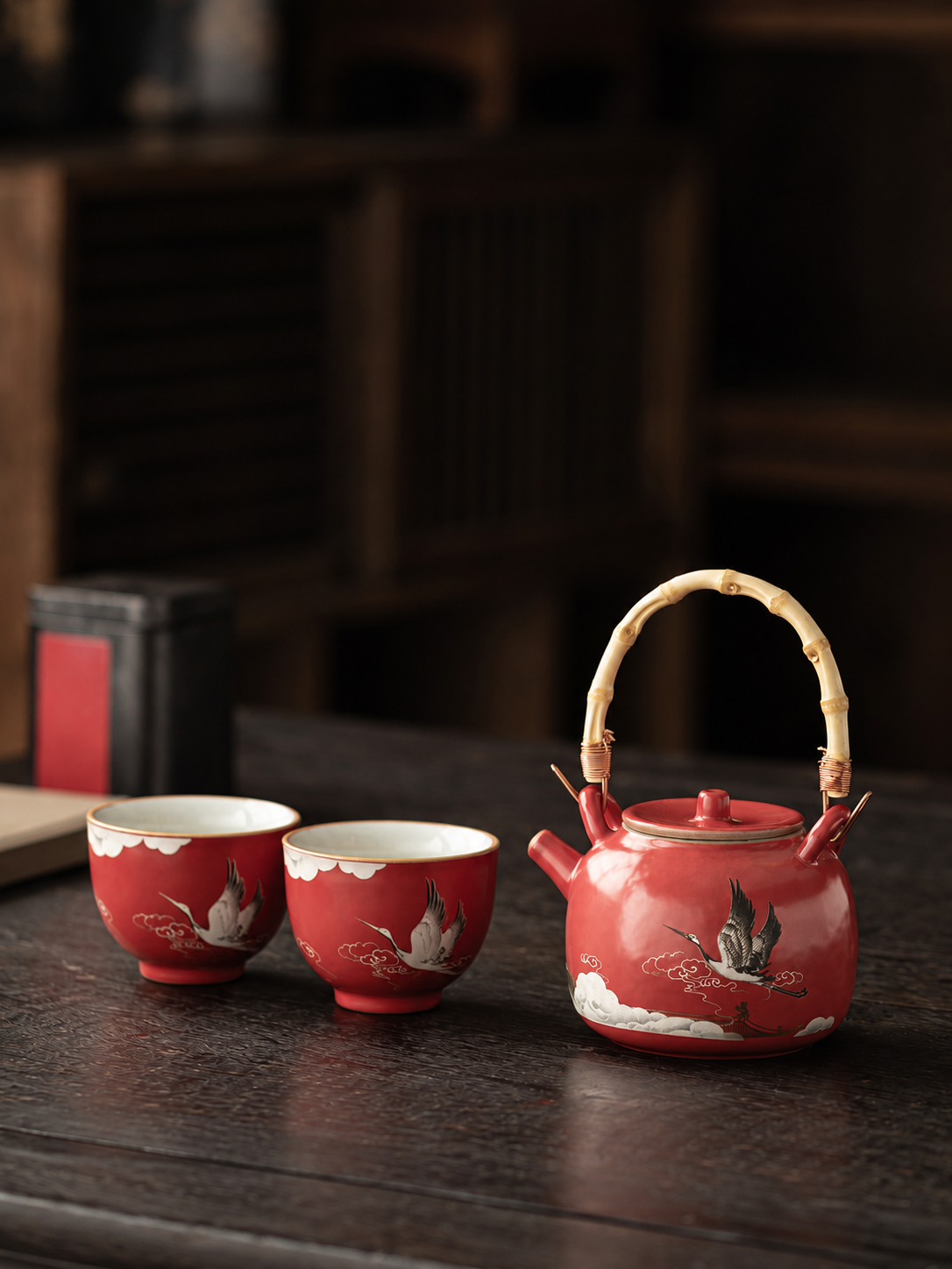 Red crane ceramic teapot and two matching cups on a dark wood table
