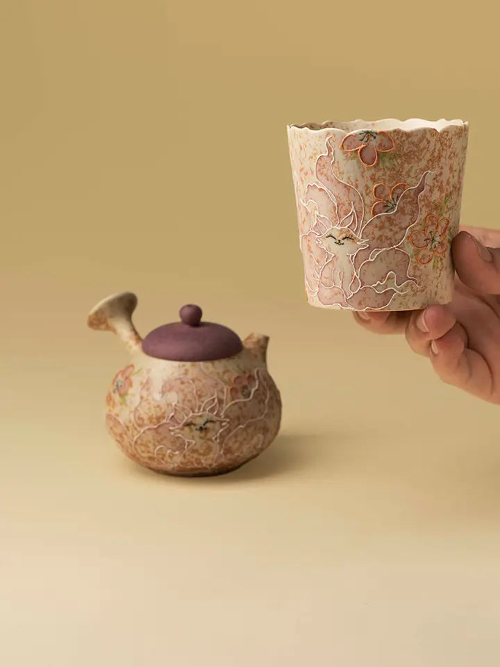 Hand holding the nine-tailed fox ceramic cup with a decorative teapot in the background
