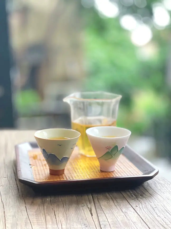 Grey-blue mountain relief and pink-green landscape teacups on a tea tray by a bright window