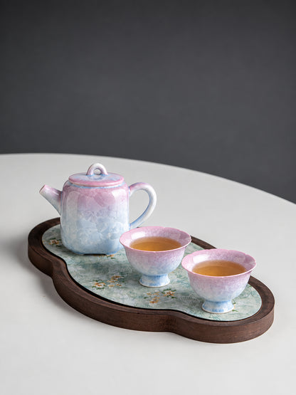 Pink-to-blue crystalline glaze teapot and two flower-shaped cups filled with tea on a wooden tea tray