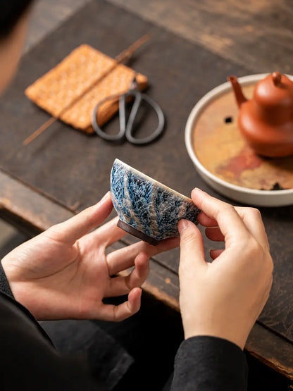 Hands holding a blue-and-white faceted ceramic tea cup beside a gongfu tea setup
