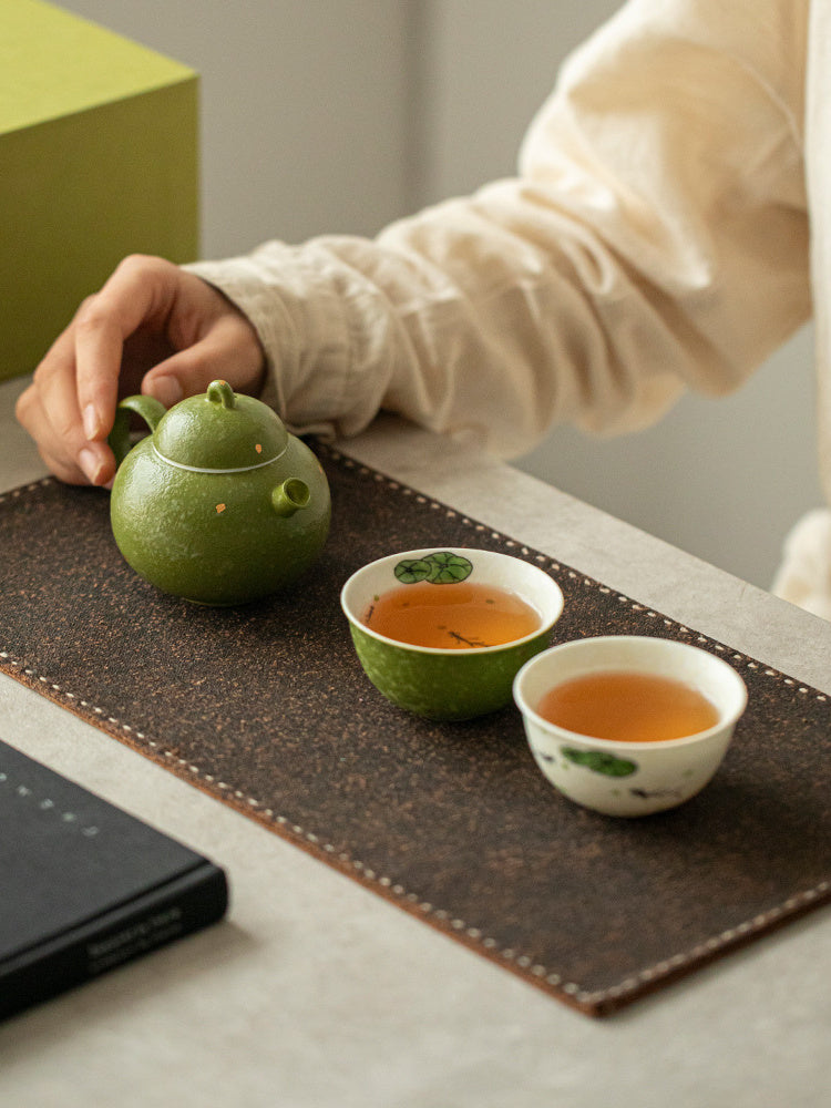 Small green ceramic teapot with green and ivory fish-motif teacups arranged on a tea mat