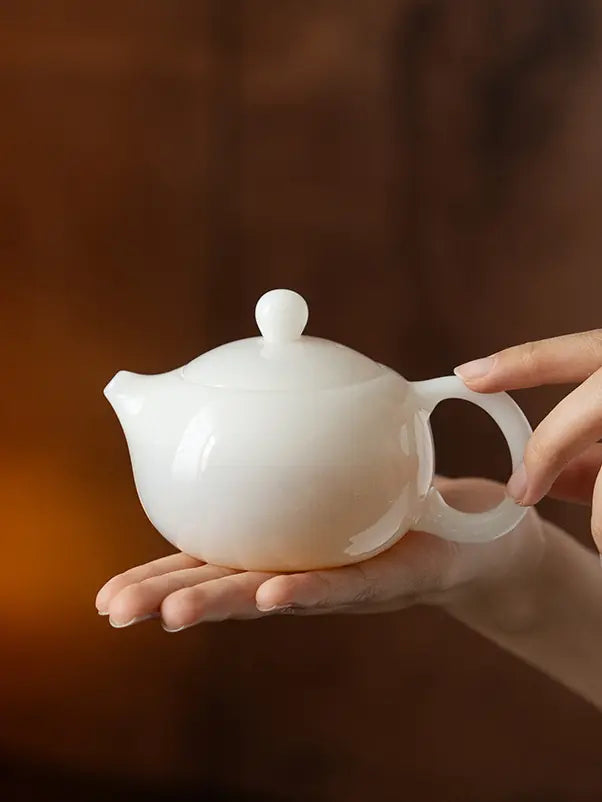 White ceramic Xishi teapot held in two hands against a warm brown background
