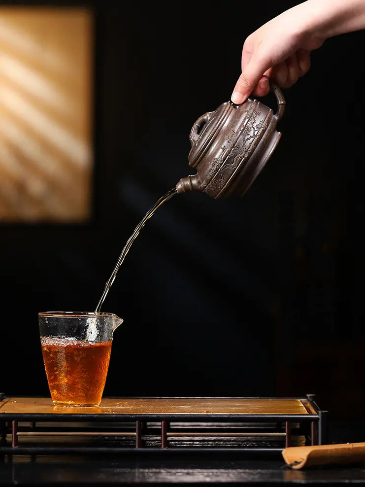 Hand pouring amber tea from a bronze-brown bamboo drum zisha teapot into a glass cup