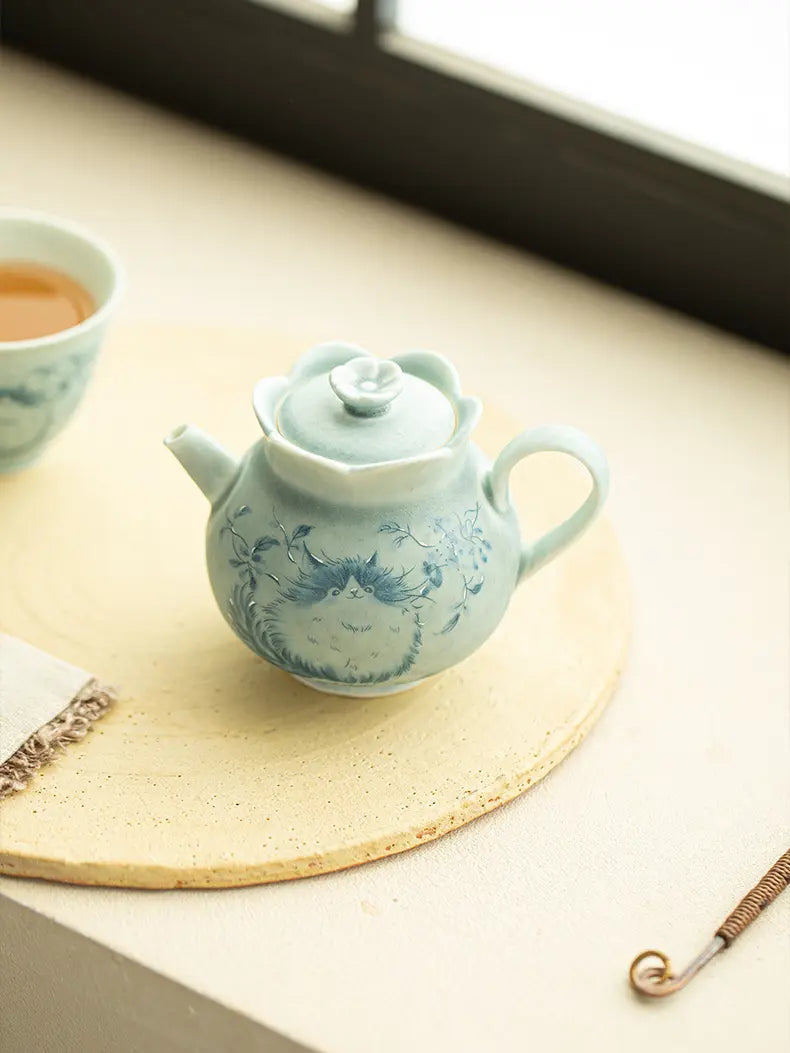 Cat-motif ceramic teapot in soft sunlight with a teacup and tea tool nearby
