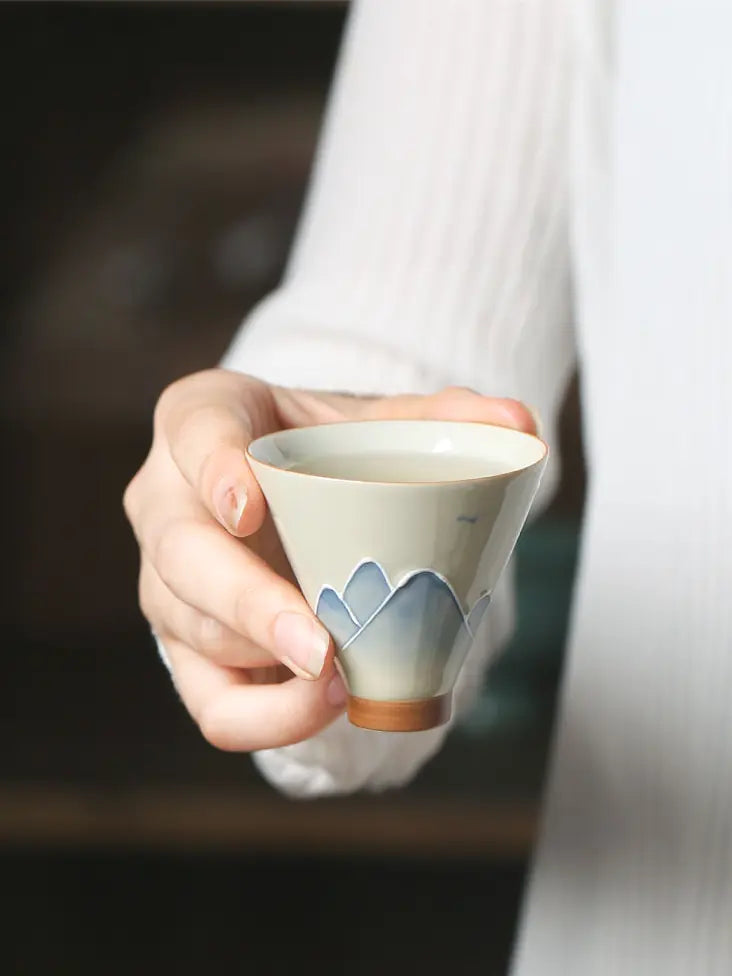 Hand holding an ivory ceramic teacup with layered grey-blue mountain relief
