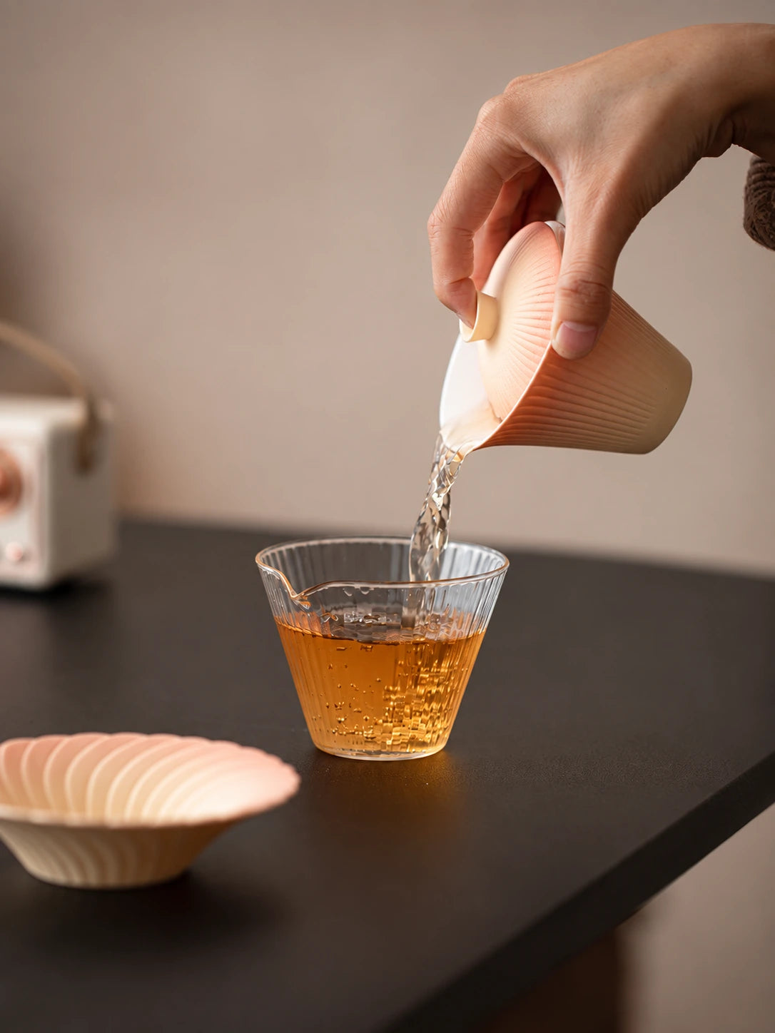 Hand pouring tea from a gradient ceramic gaiwan into a clear ribbed glass pitcher
