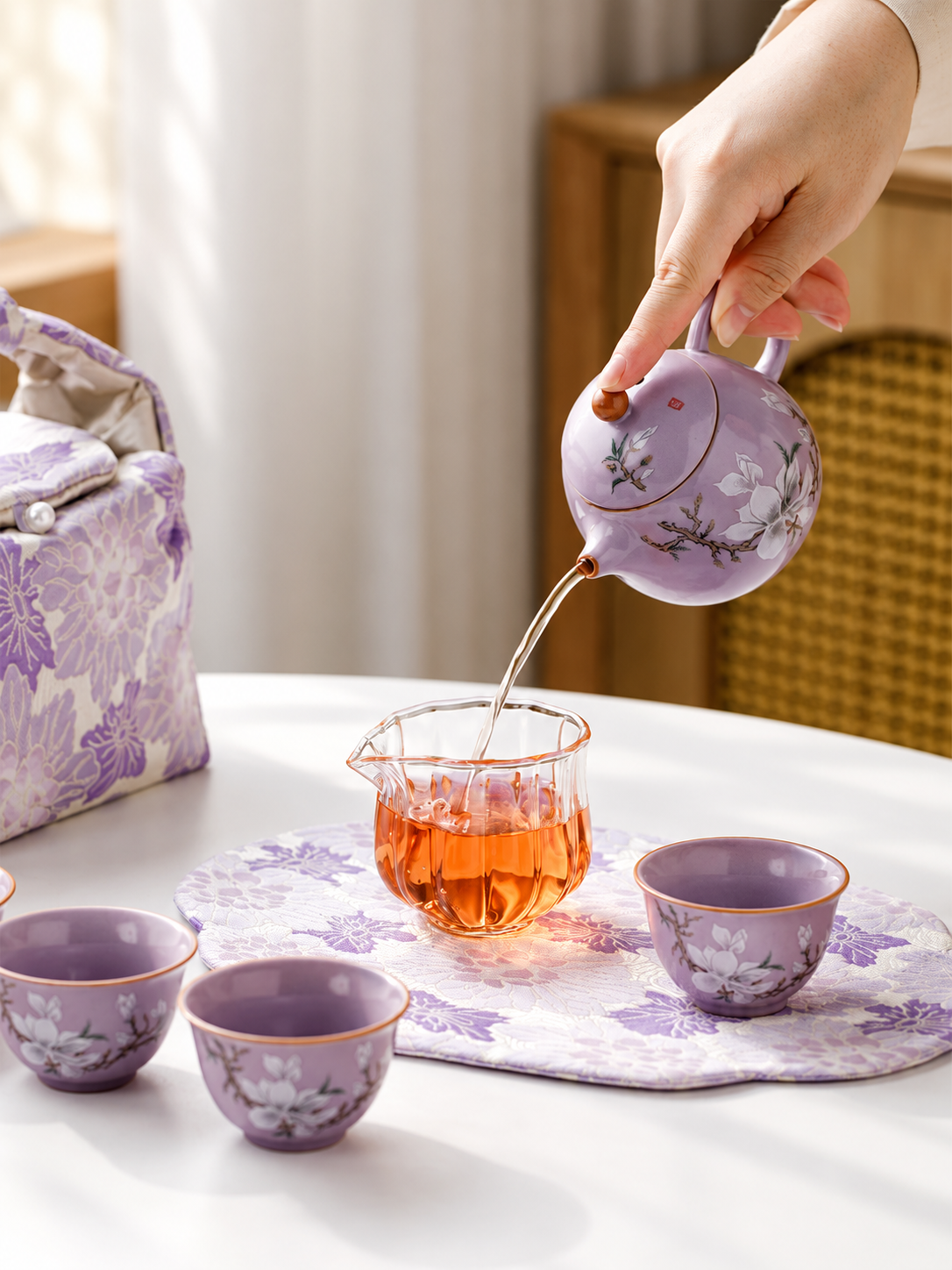 Purple magnolia teapot pouring tea into a glass server beside four matching cups
