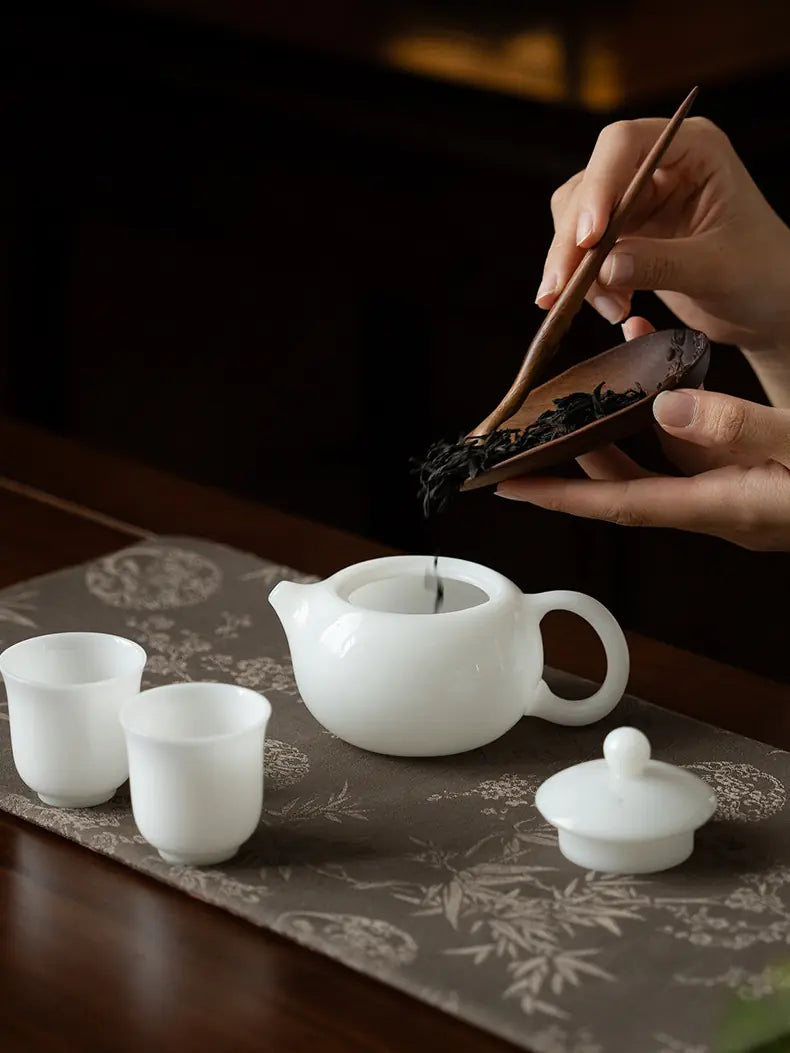 Loose tea leaves being added to a white ceramic Xishi teapot beside two cups
