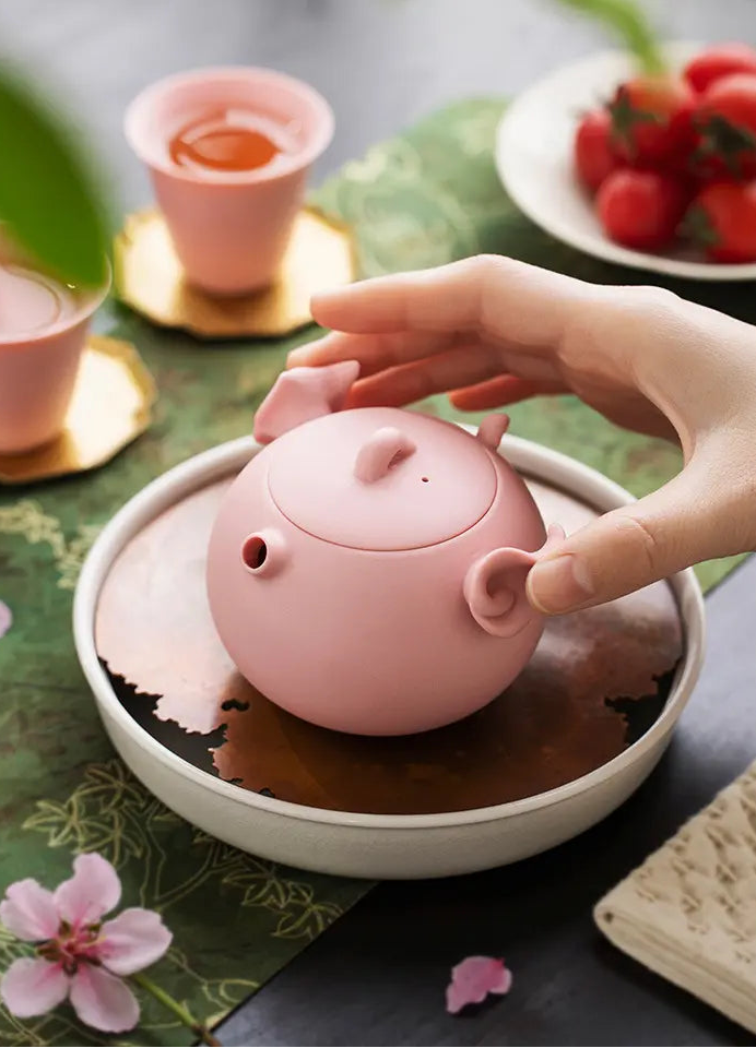 Hand reaching for a pink pig-shaped zisha teapot on a tea tray
