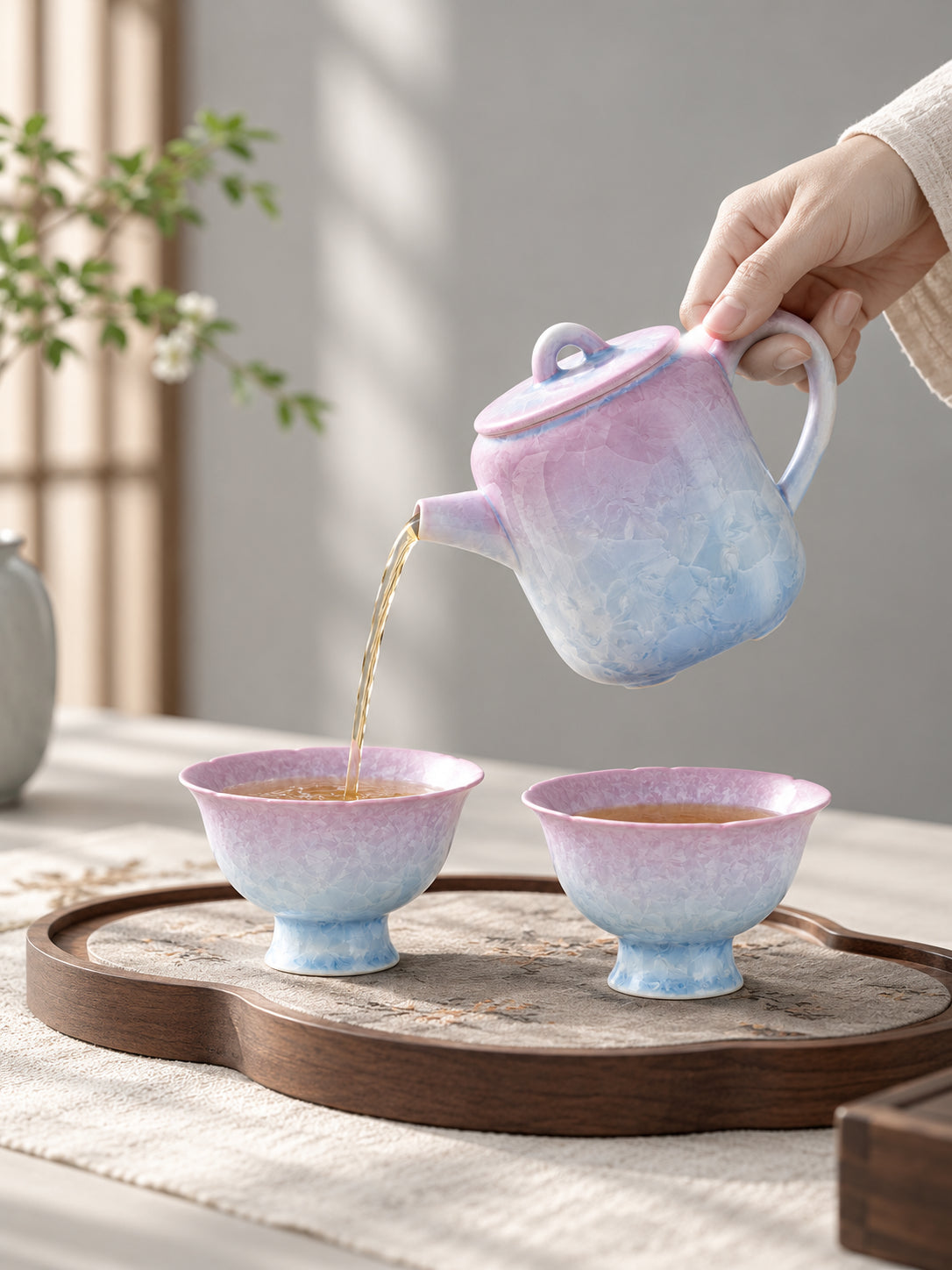 Hand pouring amber tea from a pink-to-blue crystalline glaze teapot into one of two matching flower-shaped cups