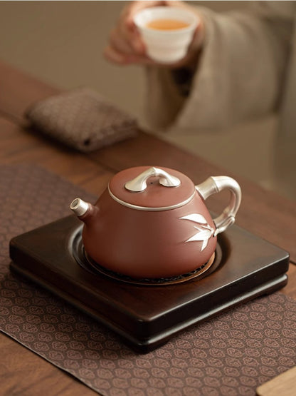 Reddish-brown Yixing zisha teapot with silver bamboo accents on a wooden tea stand