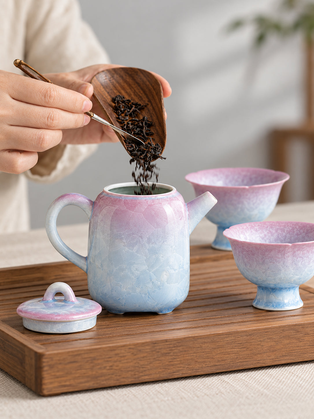 Loose tea leaves being added to an open pink-to-blue crystalline glaze teapot with two matching cups