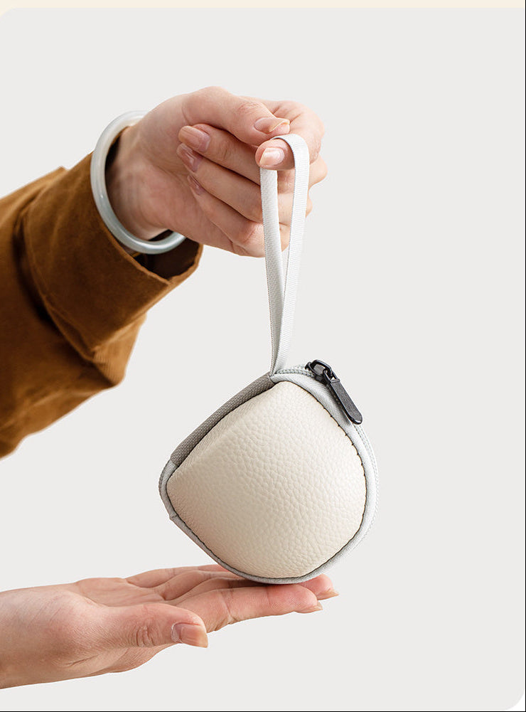 Palm-sized cream travel case with gray trim and a wrist strap held between two hands
