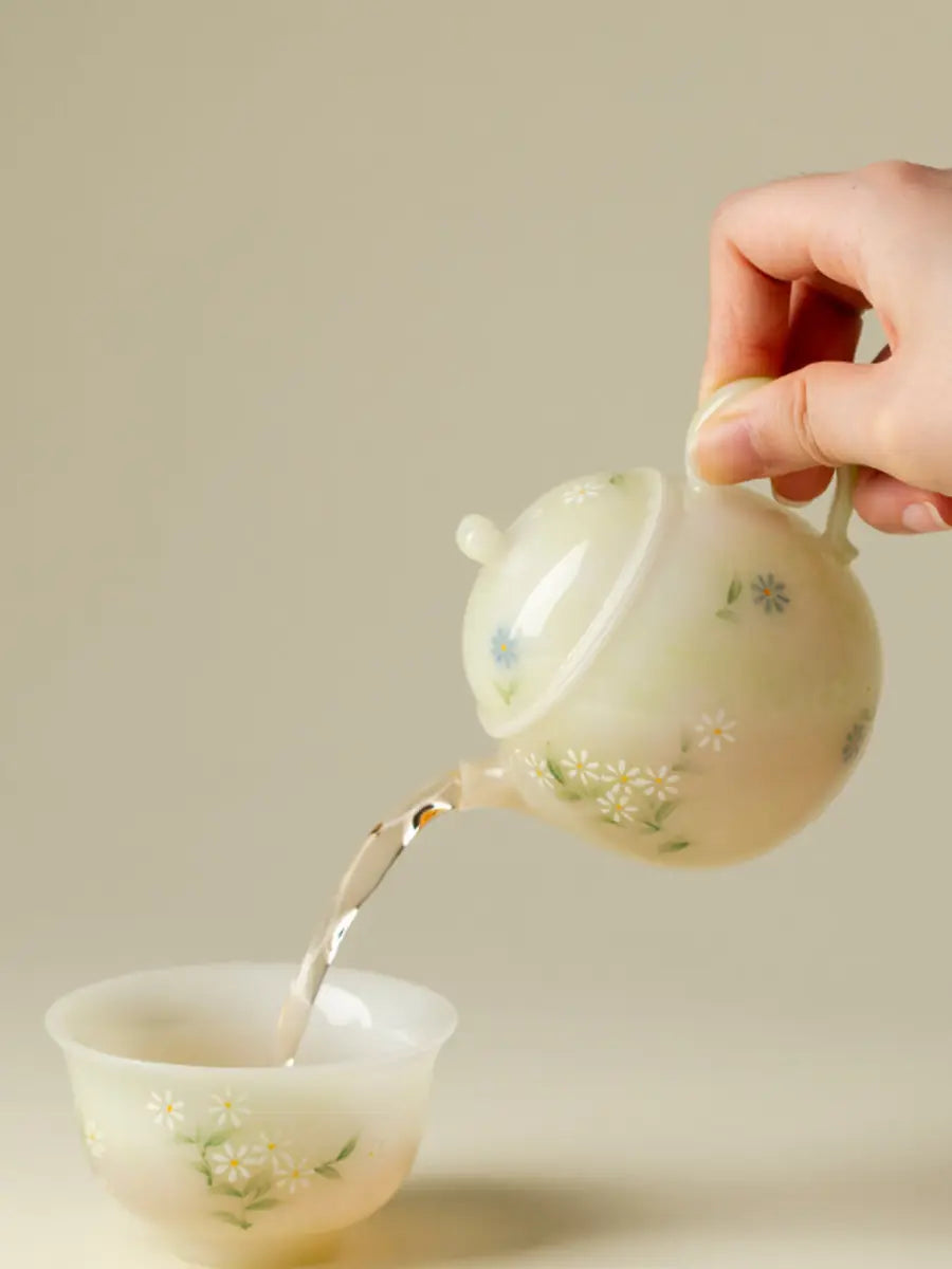 Hand pouring tea from a pale green daisy ceramic teapot into a floral cup