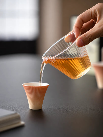 Clear ribbed glass pitcher pouring amber tea into a gradient ceramic tasting cup
