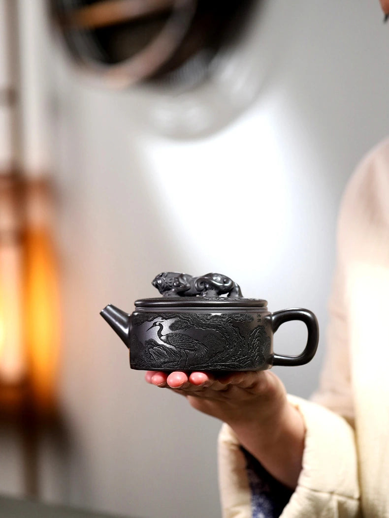Hand holding the dark carved zisha teapot to show its low Hanwa profile and rounded handle