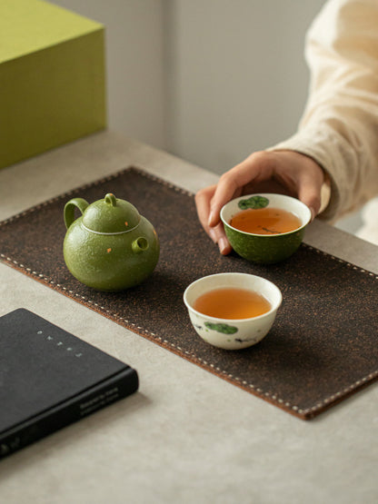 Person holding a green teacup beside the green ceramic teapot and ivory fish-motif cup