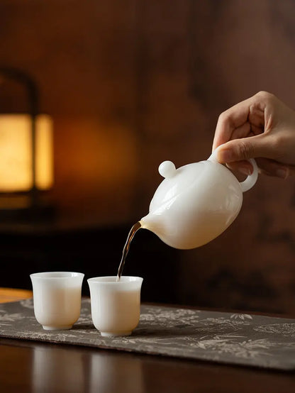 White ceramic Xishi teapot pouring amber tea into two matching cups
