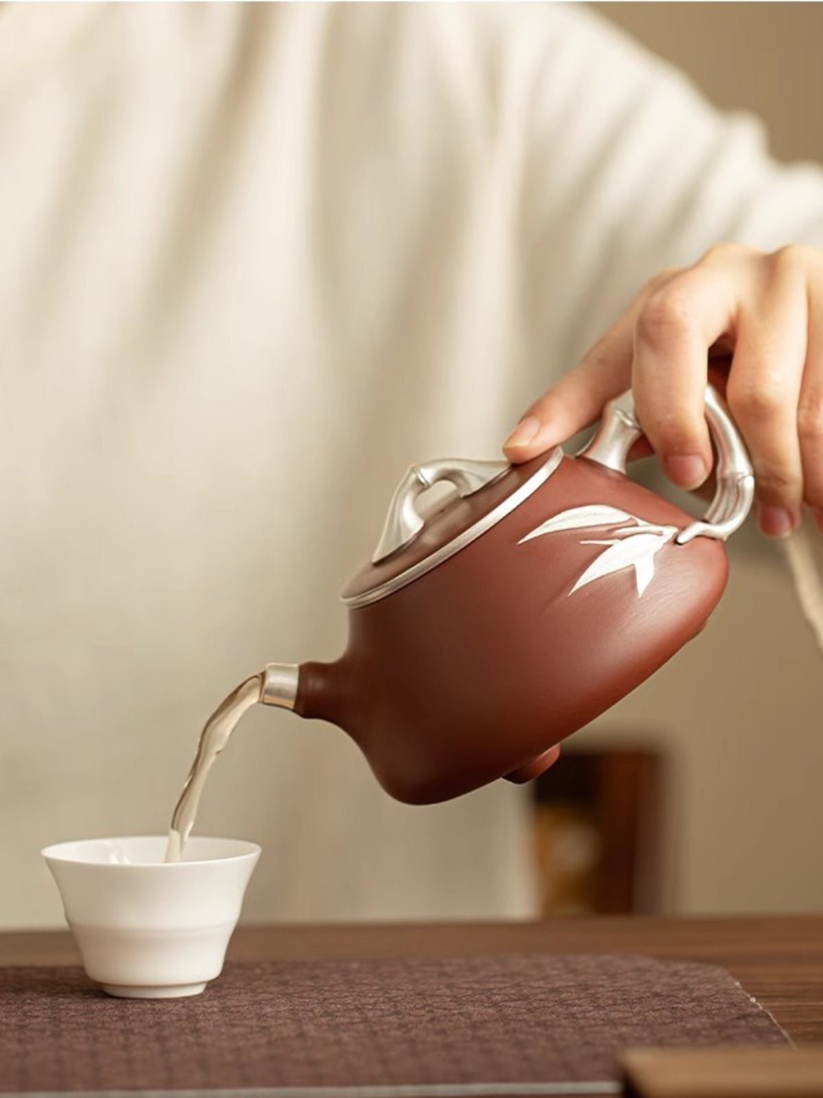 Yixing zisha teapot with silver-tone bamboo details pouring tea into a white tasting cup