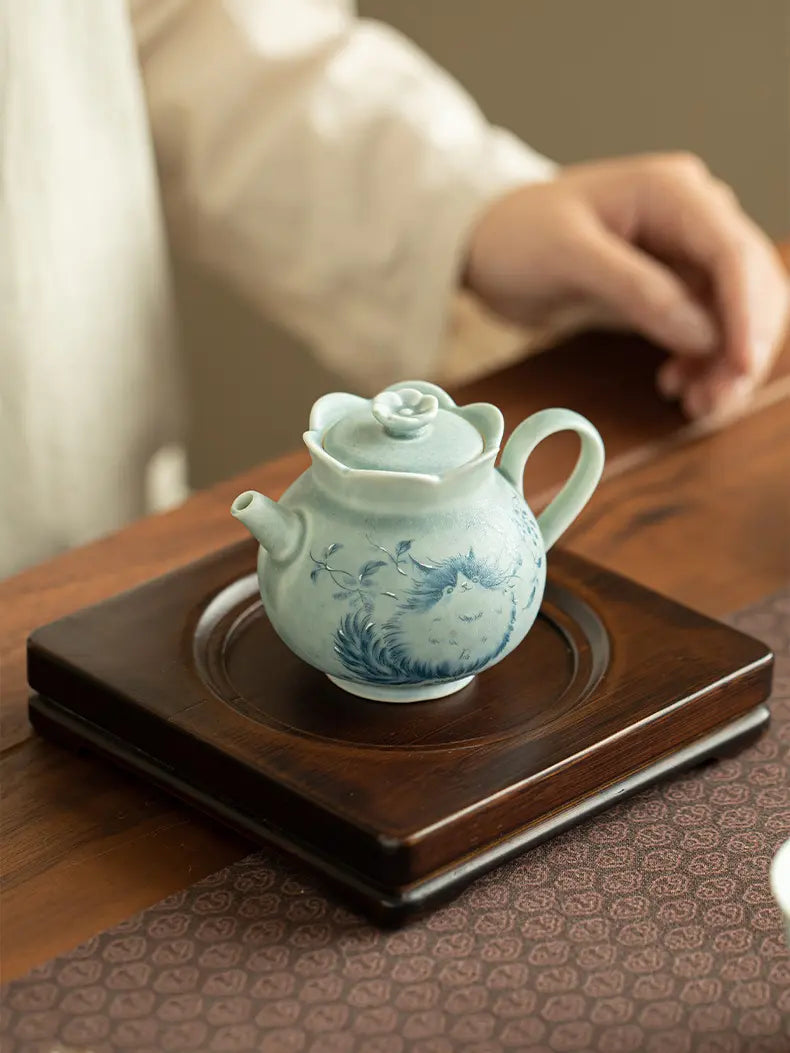 Pale green cat ceramic teapot on a wooden tea stand beside a seated person
