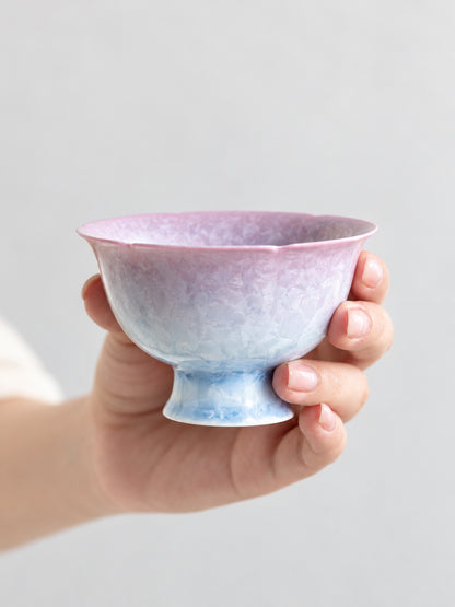 Hand holding a pink-to-blue crystalline glaze flower-shaped teacup with a pedestal foot