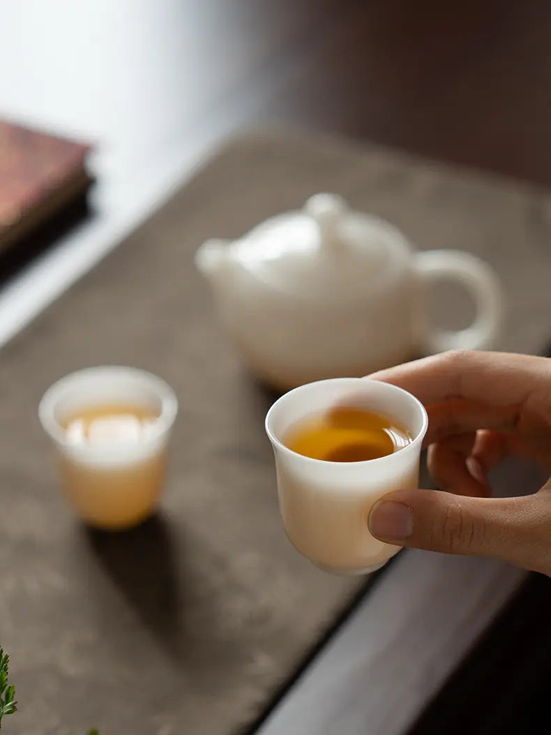 Hand holding a translucent white ceramic teacup filled with amber tea
