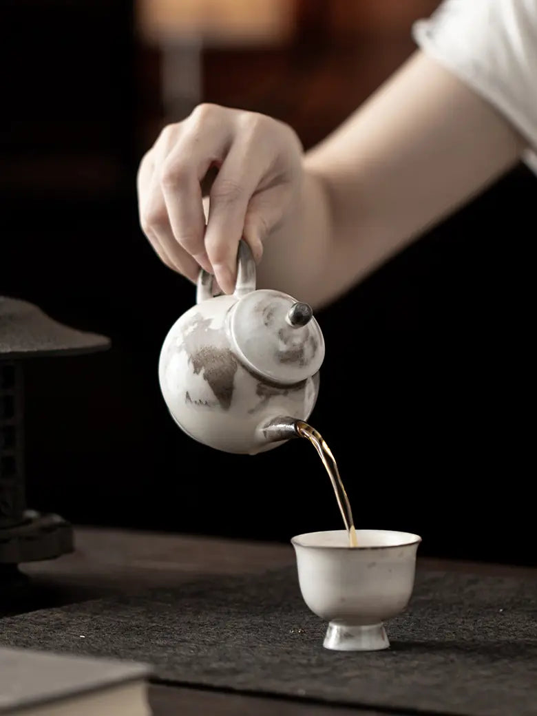 Hand pouring tea from the ink-wash ceramic teapot into a small teacup
