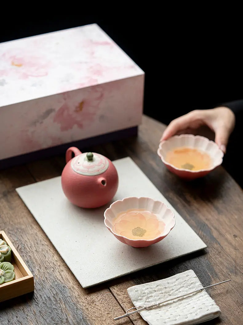 Hand lifting a flower-shaped teacup beside a pink lotus teapot and matching floral gift box
