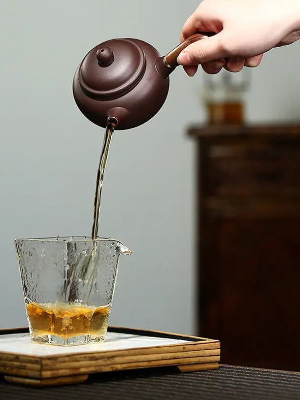 Wooden side-handle teapot pouring tea into a clear glass tea pitcher
