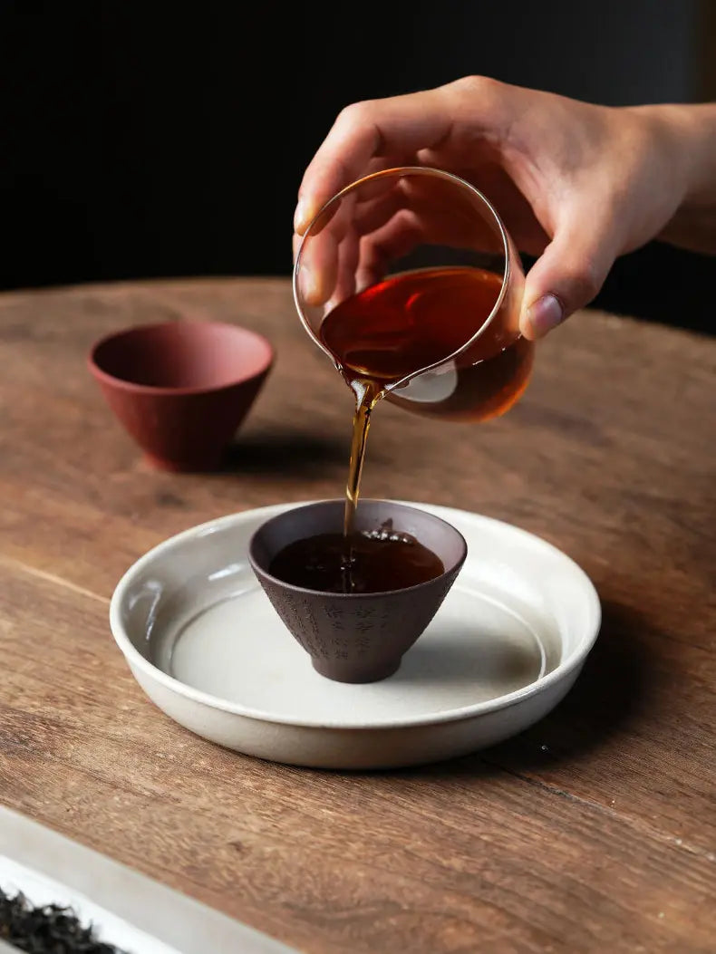 Tea poured from a glass pitcher into a dark brown Heart Sutra zisha tea cup