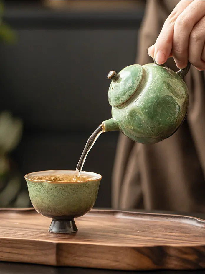 Green ceramic teapot pouring tea into a ceramic cup on a wooden tea tray
