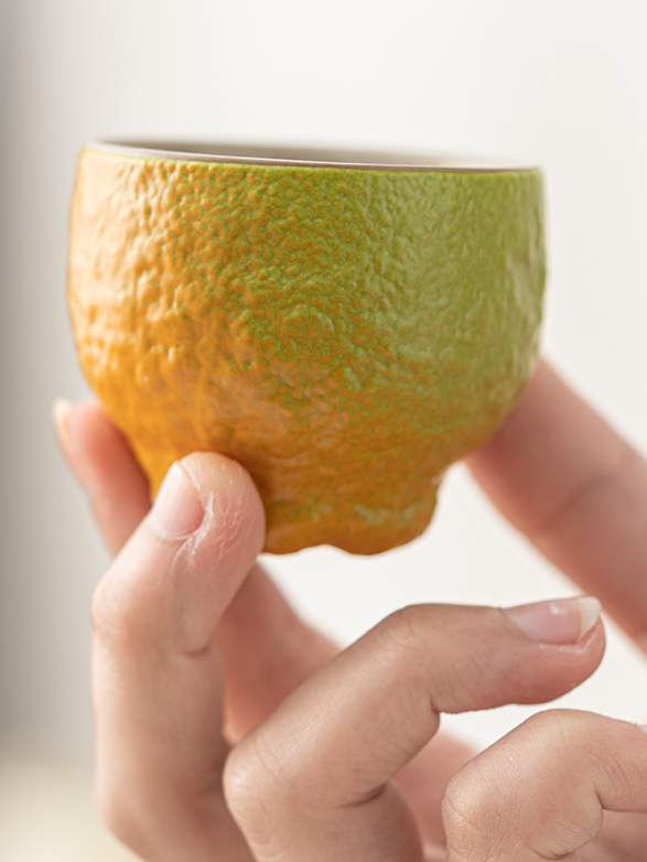 Hand holding a green-to-orange tangerine-shaped ceramic teacup with a dimpled peel texture