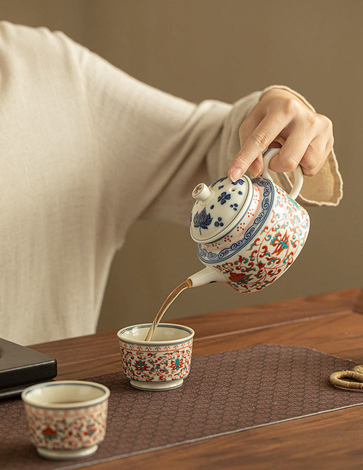 Person pouring tea from the red and green lotus teapot into a matching ceramic cup