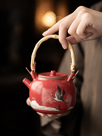Hand holding a red crane ceramic teapot by its bamboo-style arched handle

