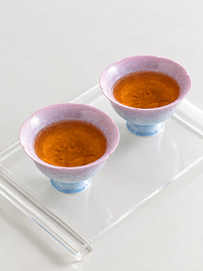 Two pink-to-blue crystalline glaze flower-shaped cups filled with amber tea on a clear tray