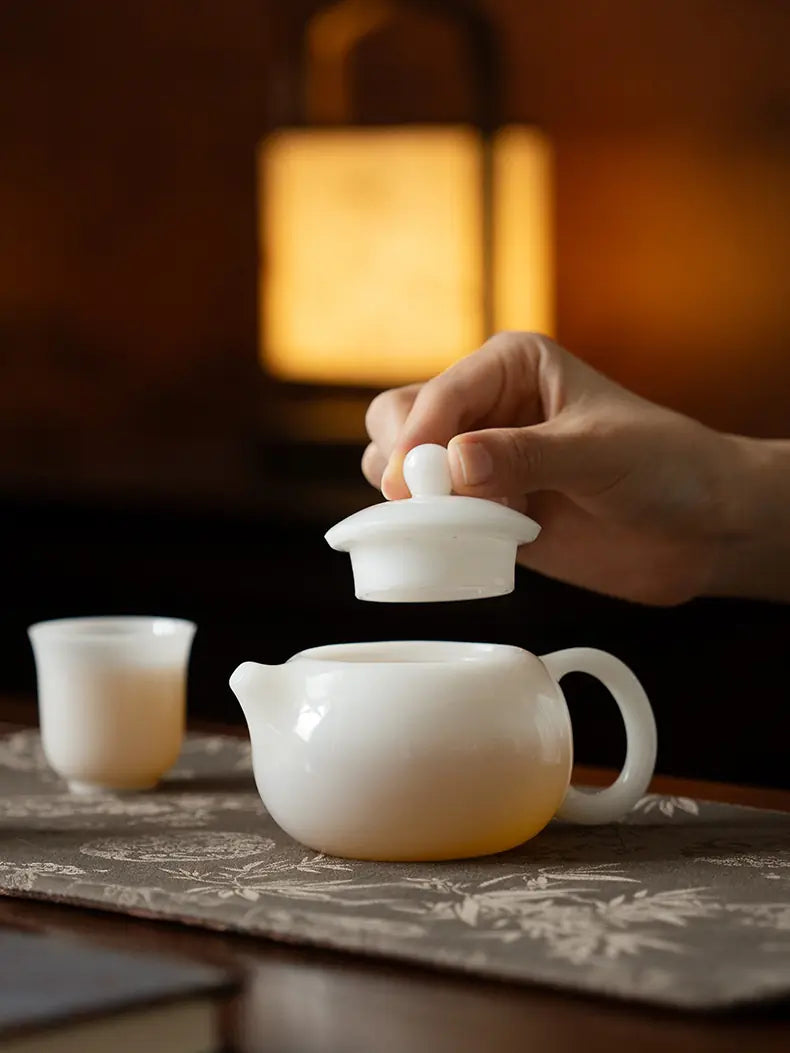 Hand lifting the lid above a white ceramic Xishi teapot with a matching cup nearby
