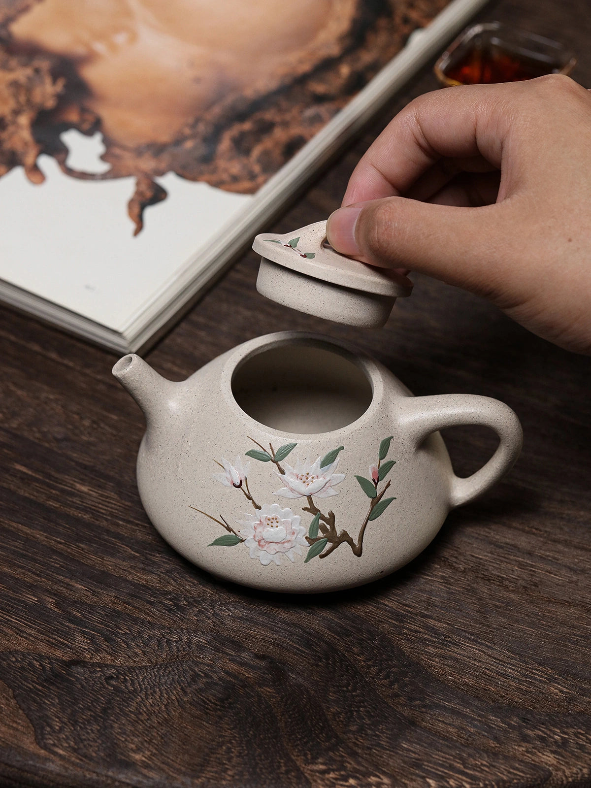 Hand lifting the lid from the floral Yixing teapot on a dark wood table