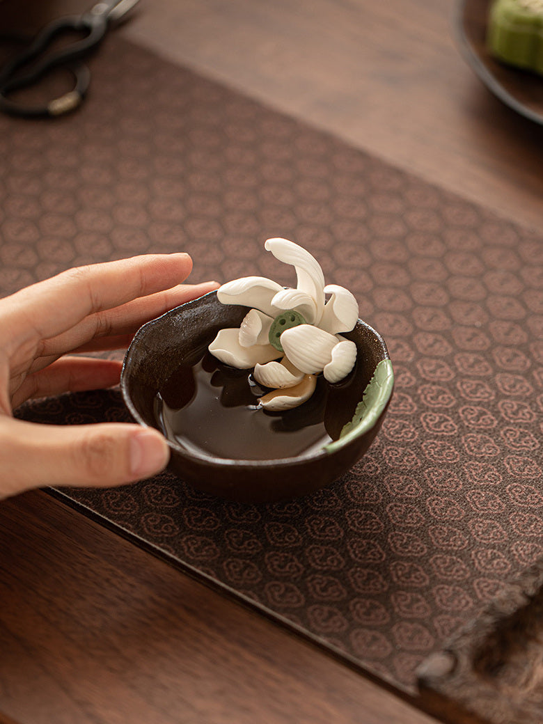 Hands arranging a wood-fired lotus tea cup on a patterned tea mat