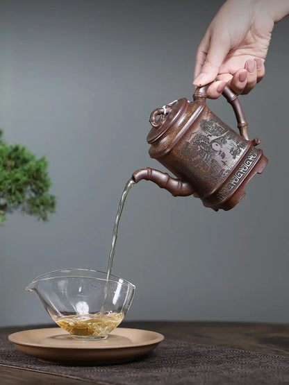 Hand pouring tea from the square bamboo-style zisha teapot into a clear glass cup
