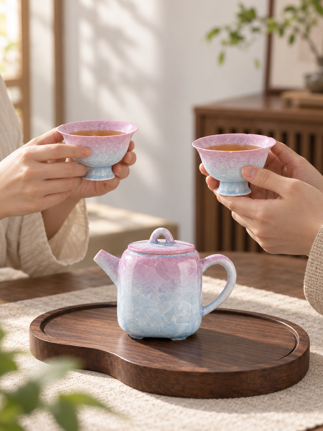Two people holding matching pink-to-blue crystalline glaze teacups beside the teapot