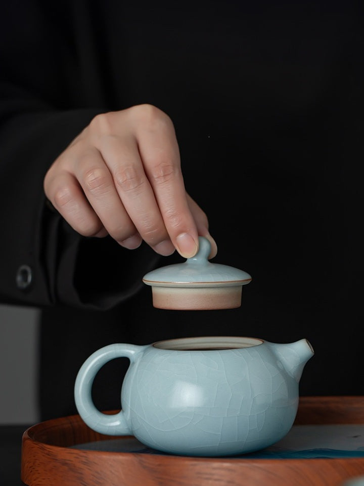 Hand lifting the fitted lid from a pale blue crackle-glazed Xishi ceramic teapot
