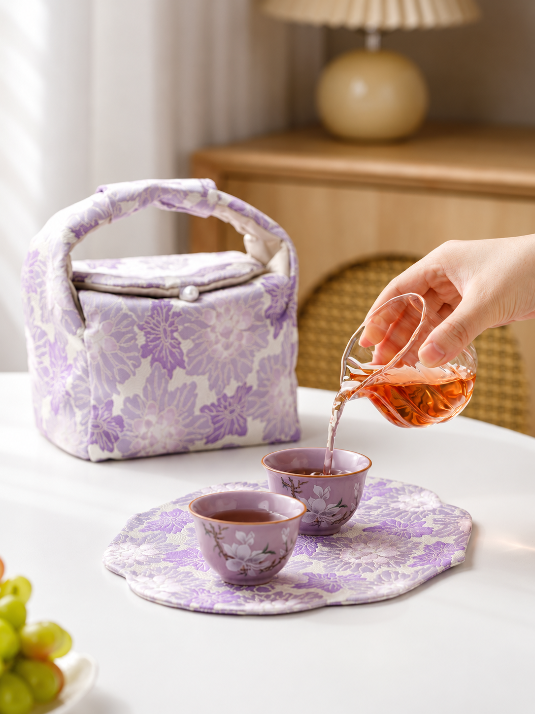 Tea poured from a glass server into purple magnolia cups beside the floral travel bag
