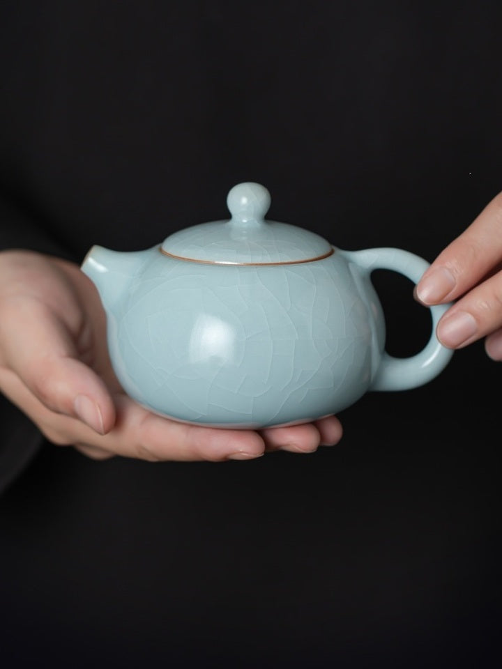 Pale blue Xishi ceramic teapot held in two hands, showing its rounded profile and balanced handle
