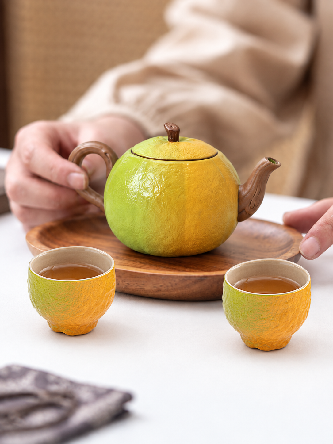 Person serving tea with a green-to-orange tangerine-shaped ceramic teapot and two matching teacups